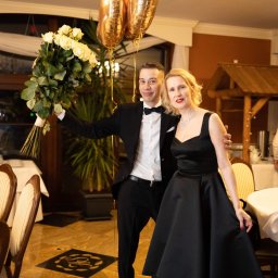 Kinga Wilda Fotografia - Elegancka para pozuje z bukietem białych róż i złotymi balonami w udekorowanym wnętrzu restauracji. Mężczyzna w smokingu, kobieta w czarnej sukni z rozkloszowanym dołem i naszyjnikiem.
