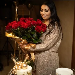 Uśmiechnięta kobieta w złotej sukience trzyma bukiet czerwonych róż z dekoracyjnym złotym papierem i wstążką, obok tort z dekoracjami w kolorze złota i czarne balony w tle.