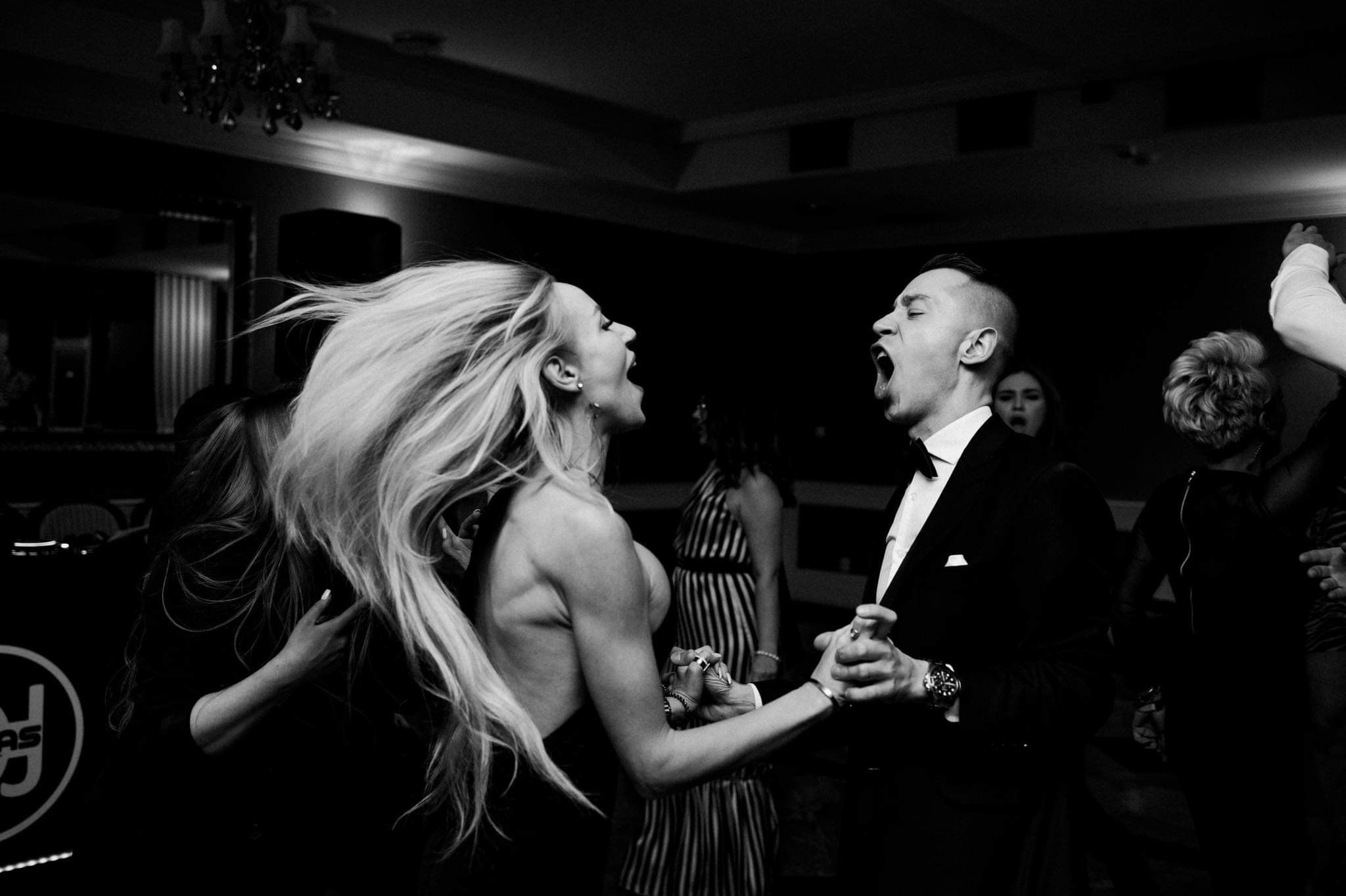 Czarno-białe zdjęcie pary młodej podczas dynamicznego tańca weselnego, oboje z otwartymi ustami w wyrazie radości, ona z rozwianymi blond włosami, on w klasycznym smokingu z muszką, w tle inni goście.