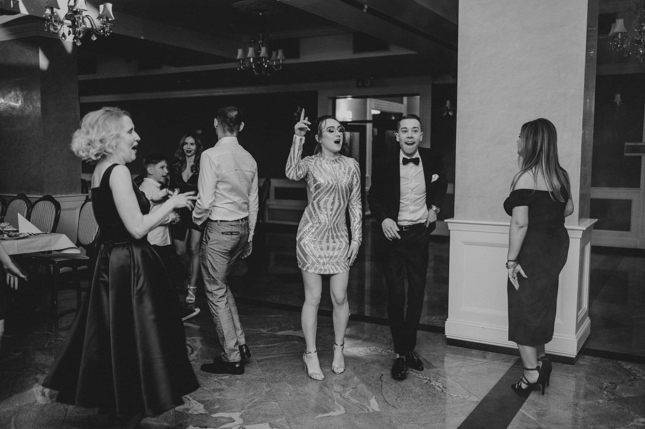 Czarno-białe zdjęcie z uroczystości: kobieta w cekinowej sukience z uniesioną ręką, mężczyzna w smokingu z zabawną miną, parkiet z marmurowym wzorem, w tle goście.