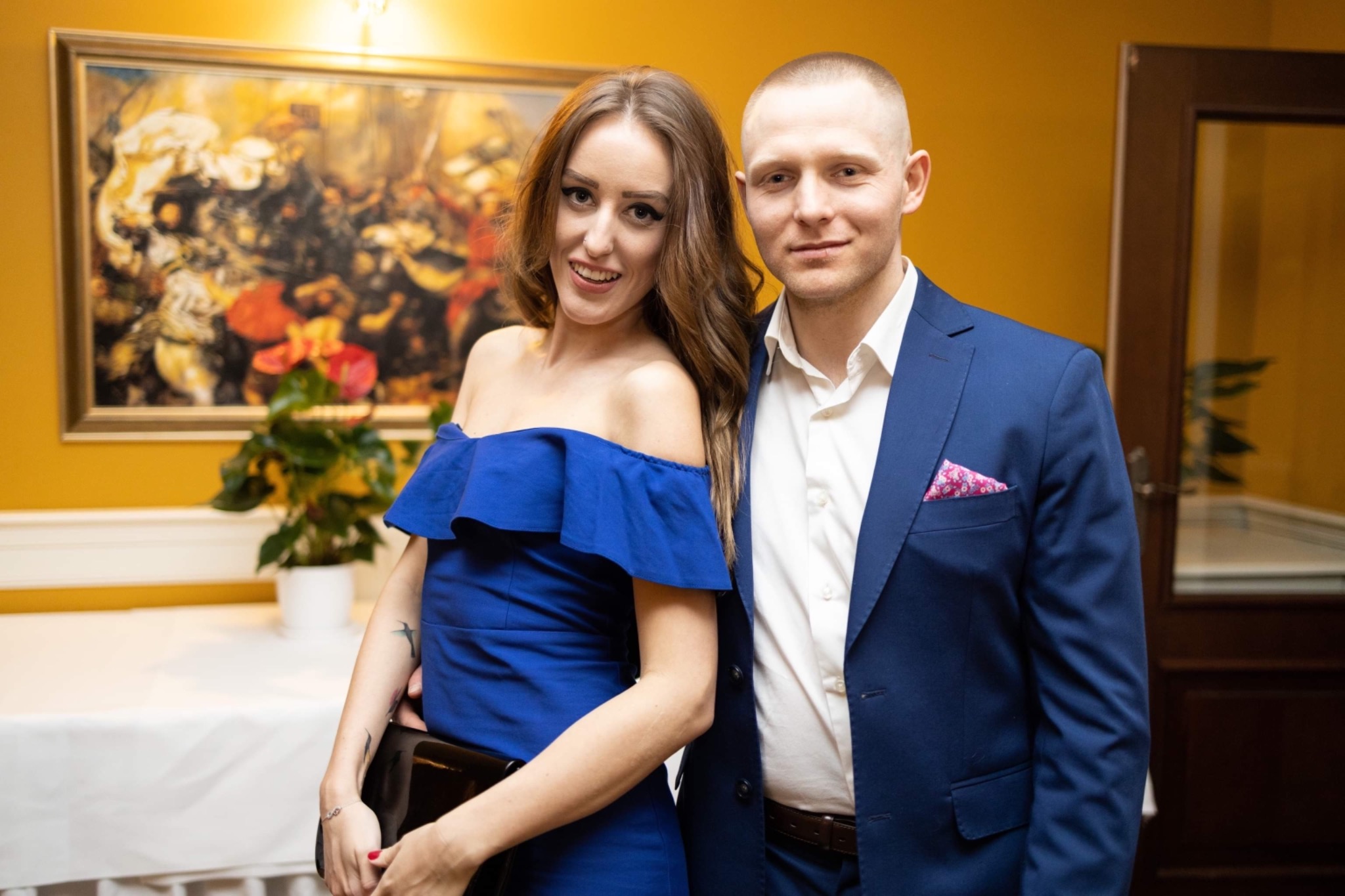 Elegancko ubrana para pozuje na tle żółtej ściany z obrazem, kobieta w niebieskiej sukience z odkrytymi ramionami trzyma czarną kopertówkę, mężczyzna w granatowym garniturze z różową poszetką.