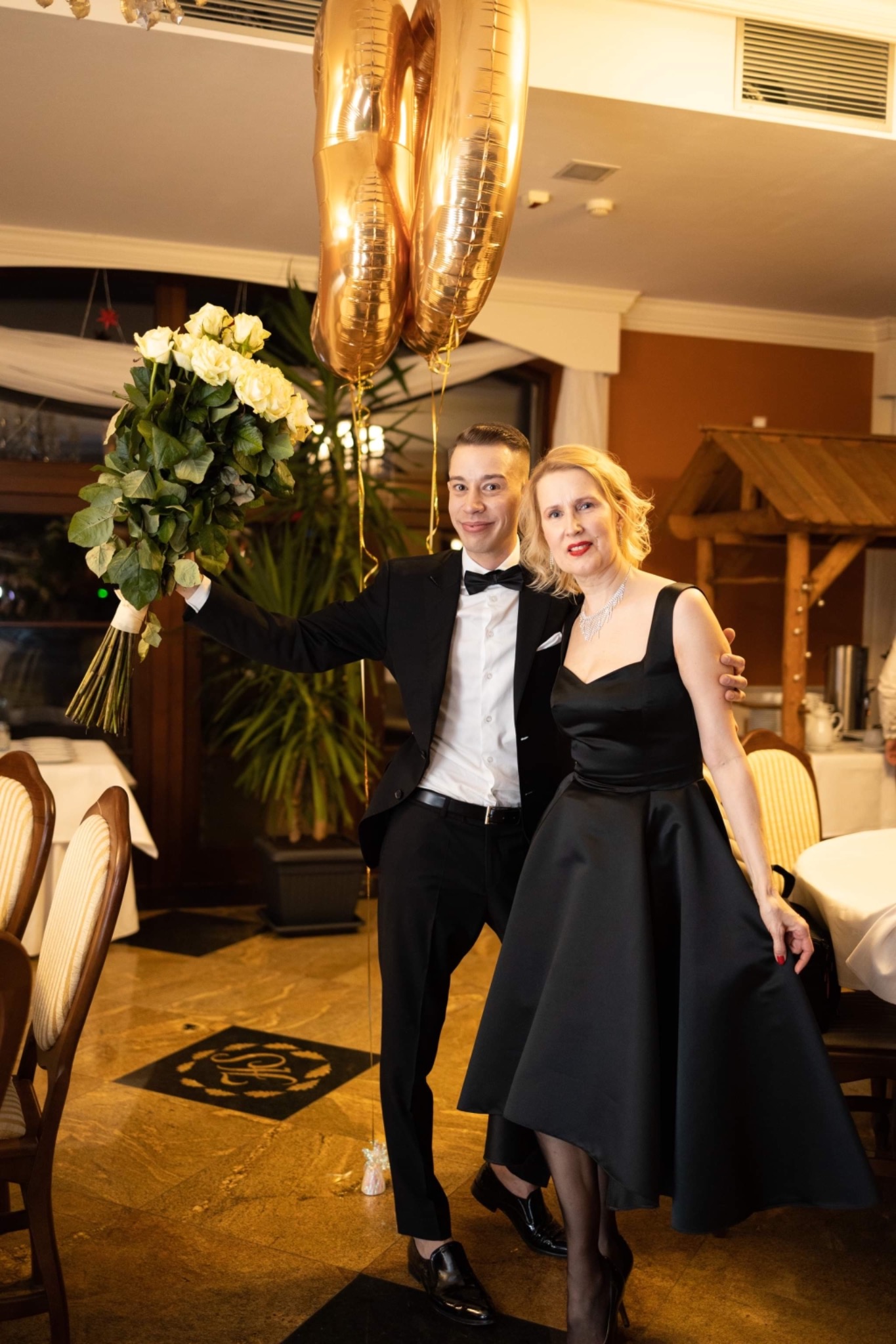 Elegancka para pozuje z bukietem białych róż i złotymi balonami w udekorowanym wnętrzu restauracji. Mężczyzna w smokingu, kobieta w czarnej sukni z rozkloszowanym dołem i naszyjnikiem.