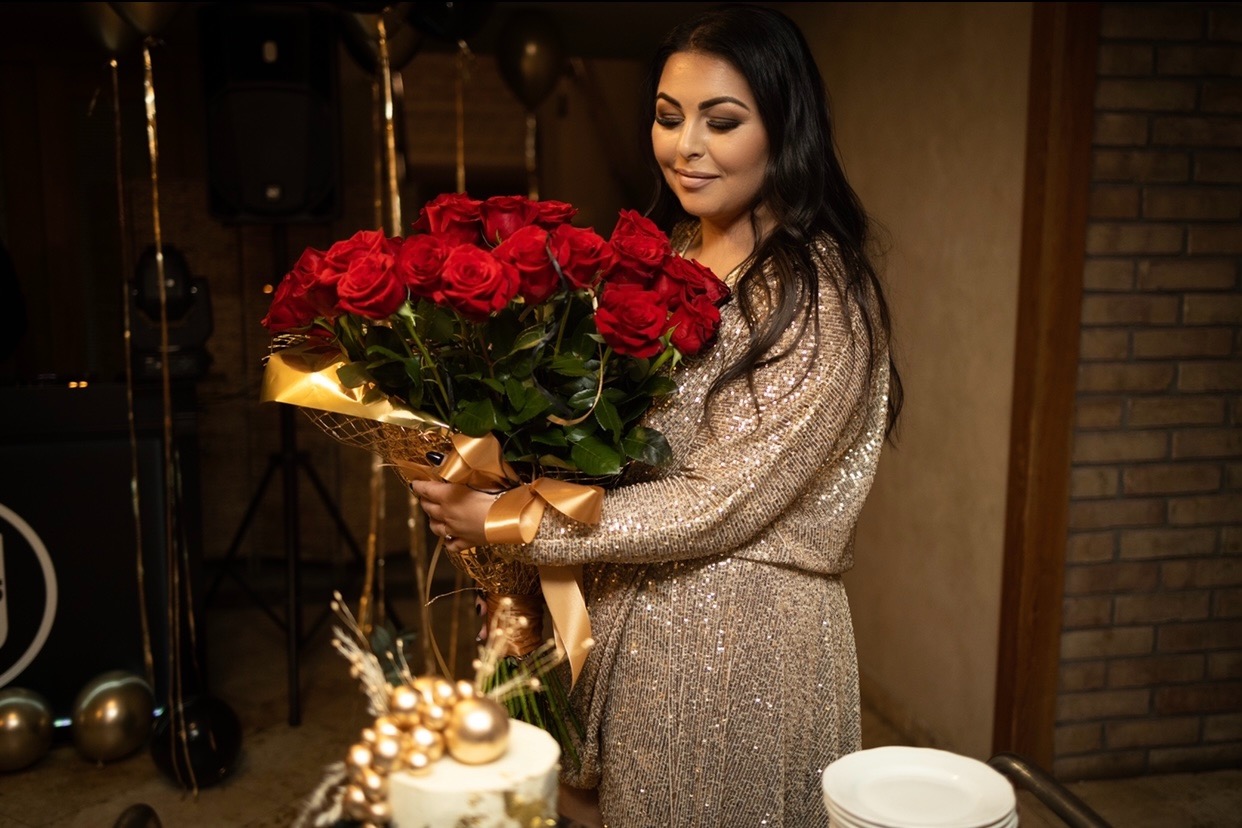 Uśmiechnięta kobieta w złotej sukience trzyma bukiet czerwonych róż z dekoracyjnym złotym papierem i wstążką, obok tort z dekoracjami w kolorze złota i czarne balony w tle.