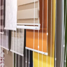 Ekspozycja różnokolorowych rolet okiennych typu dzień i noc, z białymi łańcuszkami, prezentowanych obok siebie na białej ścianie, z fragmentem wzorzystej tapety w tle.