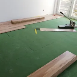 Układanie paneli podłogowych: widoczne deski, zielony podkład, narzędzie do cięcia, poziomica i okno w tle.