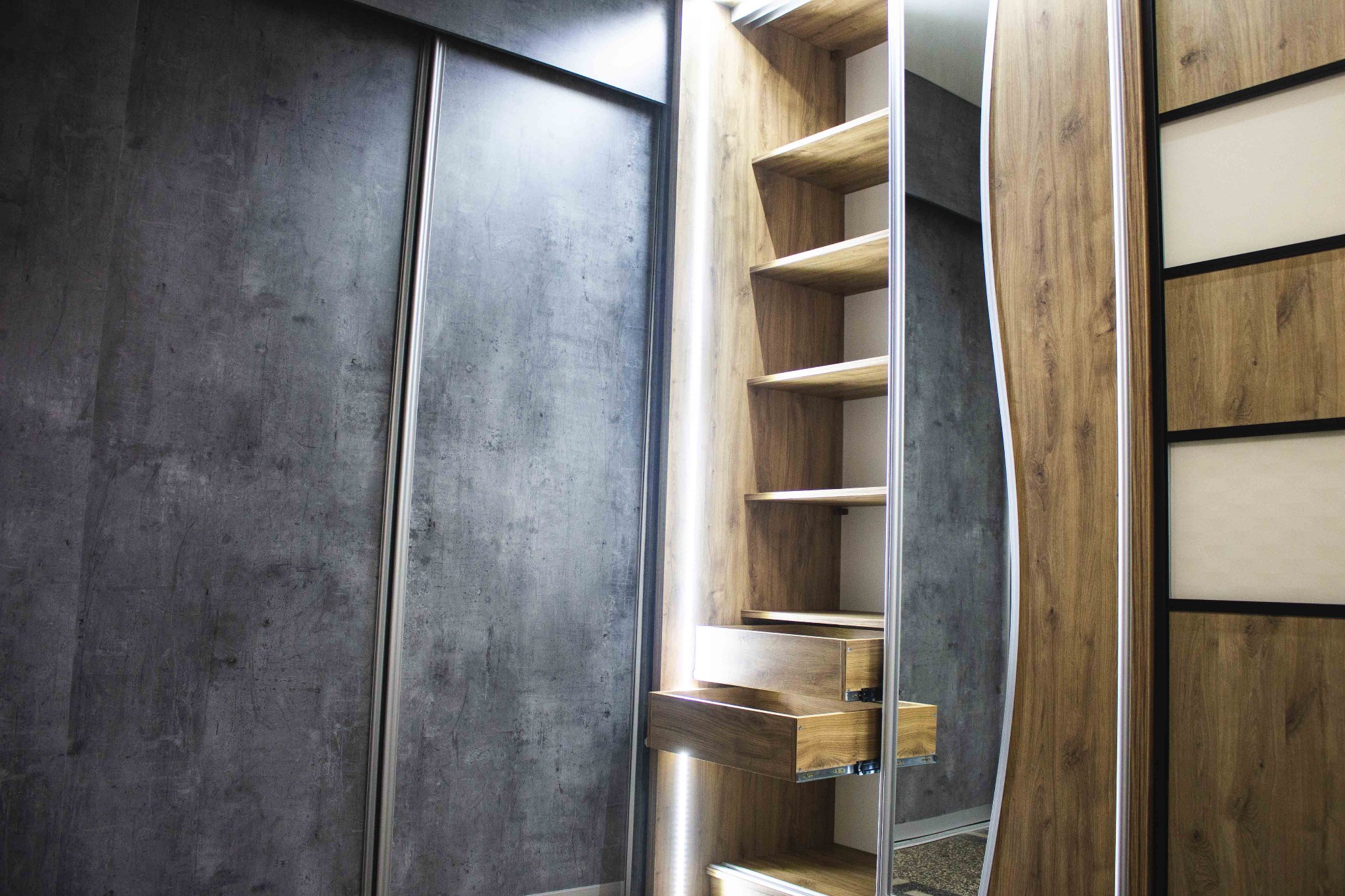 Nowoczesna szafa wnękowa z przesuwnymi drzwiami: szara, betonowa faktura frontów kontrastuje z ciepłym odcieniem drewna wewnątrz, podświetlona listwami LED, z wysuwanymi szufladami.