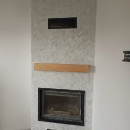 Obudowa kominka wyłożona jasnoszarą mozaiką w jodełkę, z drewnianą półką i czarnym wkładem kominkowym.