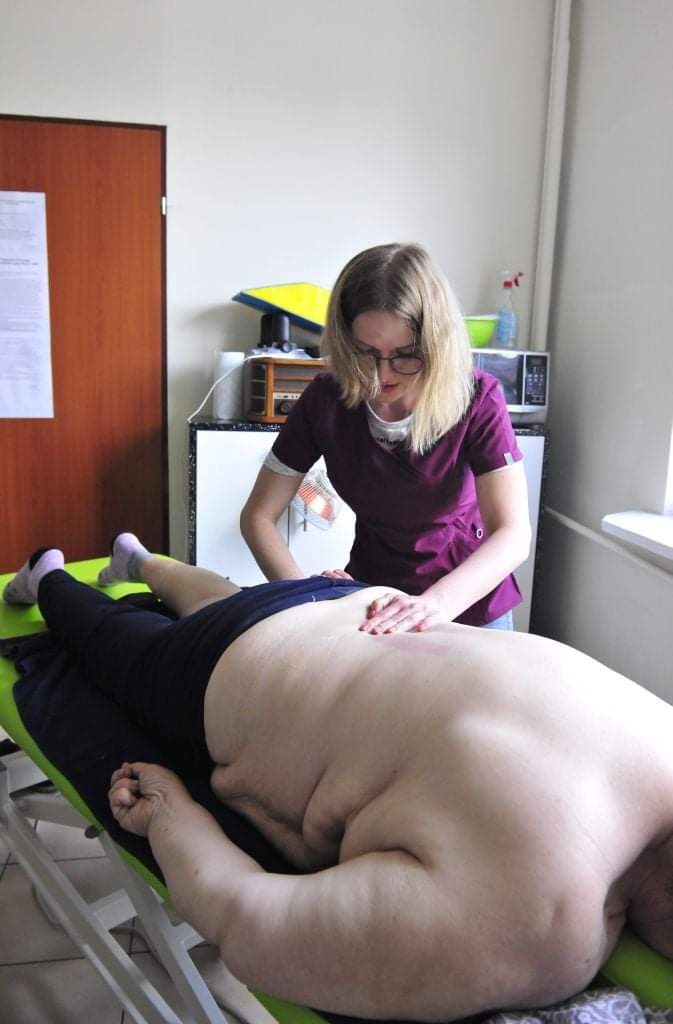 Kobieta w fioletowym uniformie wykonuje masaż pleców pacjenta leżącego na zielonym łóżku rehabilitacyjnym w gabinecie.