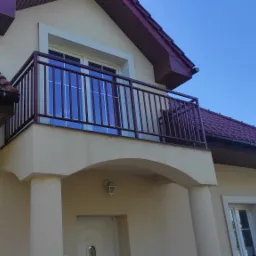Balkon z bordową metalową balustradą w nowoczesnym domu z beżową elewacją i czerwoną dachówką, widok z dołu na tle błękitnego nieba.