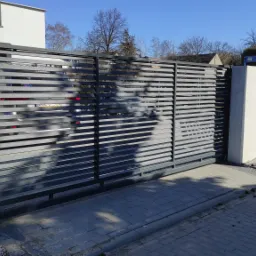Nowoczesna, przesuwna brama posesyjna z poziomych, szarych profili, widok z zewnątrz na tle białego muru i ulicy z kostki brukowej.