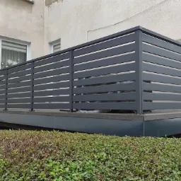 Szara, pozioma balustrada balkonowa z metalowych profili, widok z poziomu krzewów, elewacja budynku w tle.