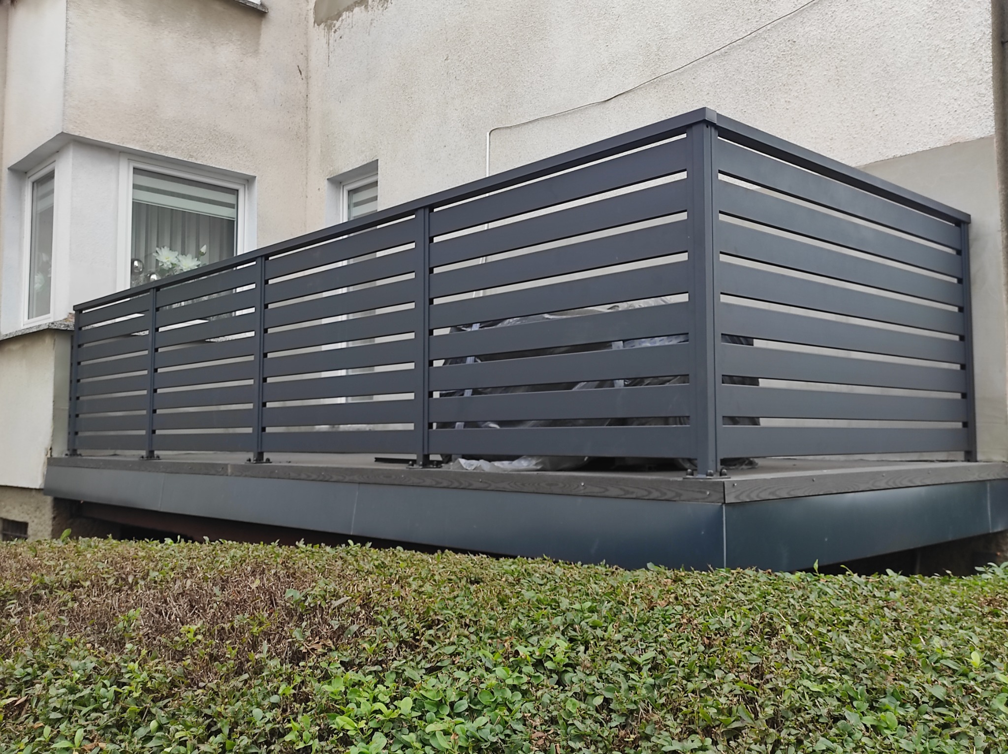 Szara, pozioma balustrada balkonowa z metalowych profili, widok z poziomu krzewów, elewacja budynku w tle.