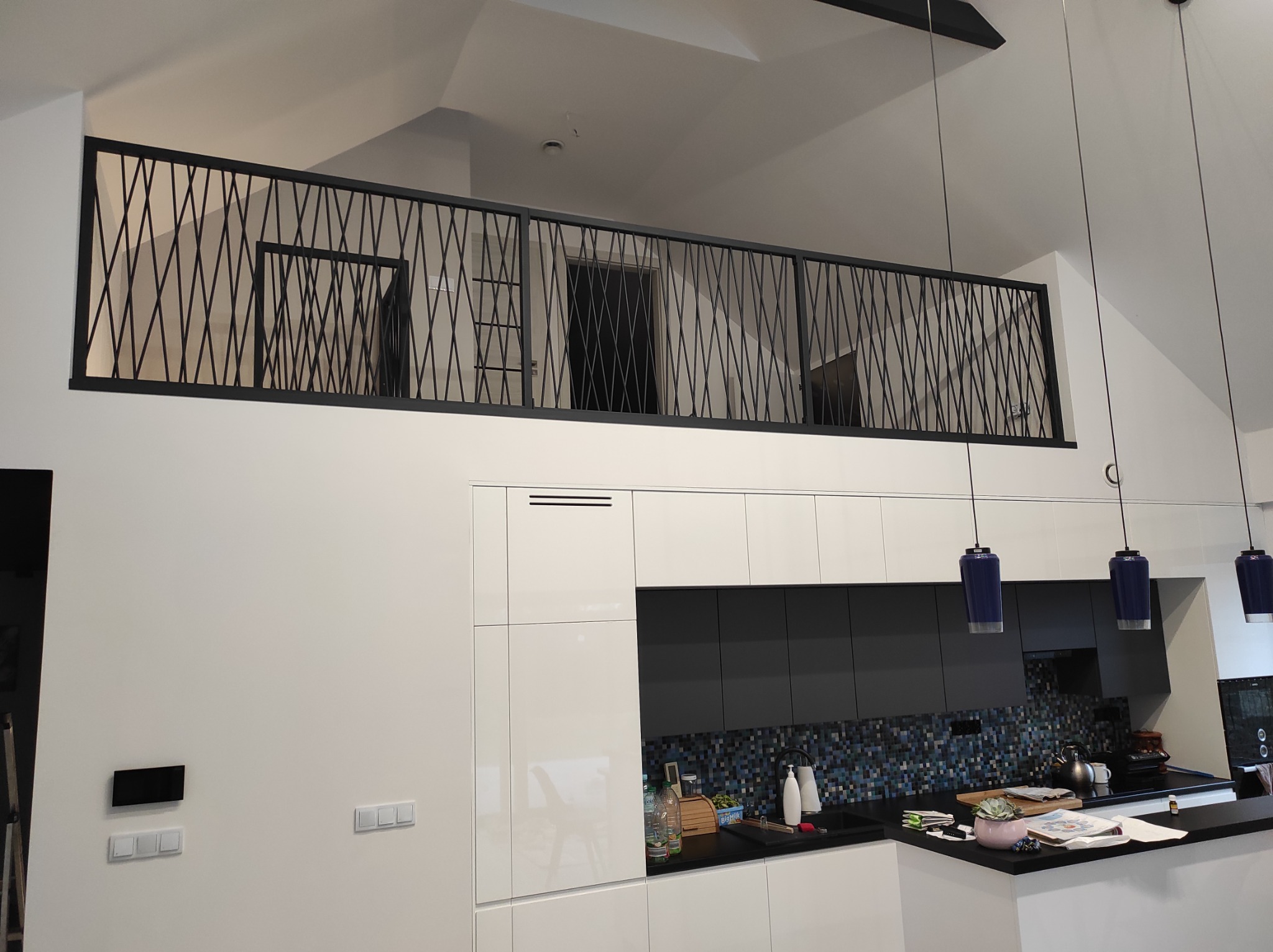 Nowoczesna balustrada metalowa z czarnych prętów krzyżujących się na antresoli w jasnym wnętrzu domu z widokiem na kuchnię.