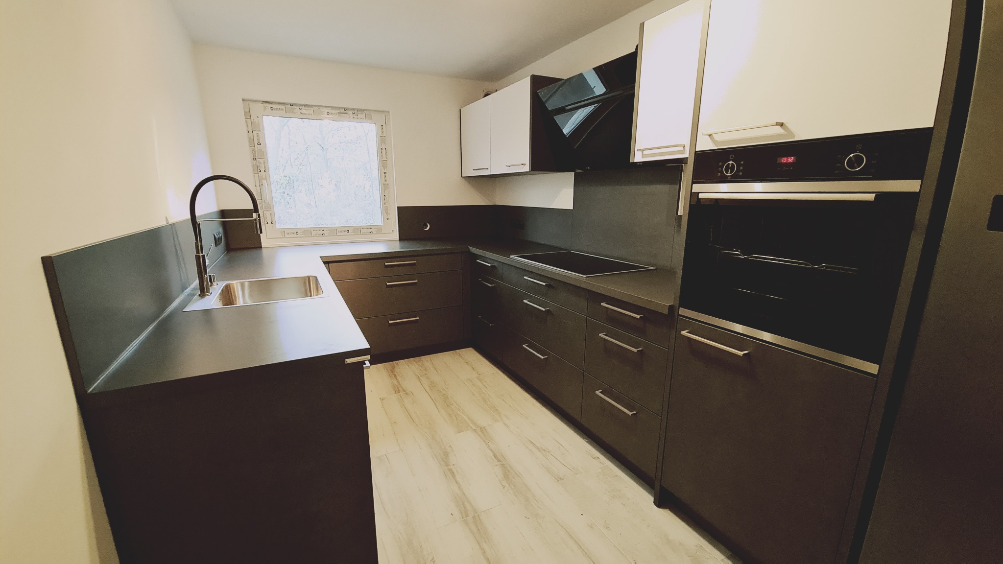 Nowoczesna kuchnia z ciemnymi, matowymi szafkami, jasnymi blatami i drewnopodobną podłogą, widok na okno z widokiem na drzewa.