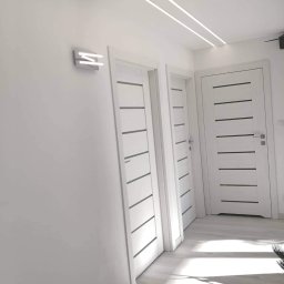 NATI&DOM remonty- wykończenia wnętrz Izabela Polak - Wnętrze w bieli: trzy białe drzwi z poziomymi, ciemnymi wstawkami, oświetlenie sufitowe LED i nowoczesny kinkiet na ścianie.