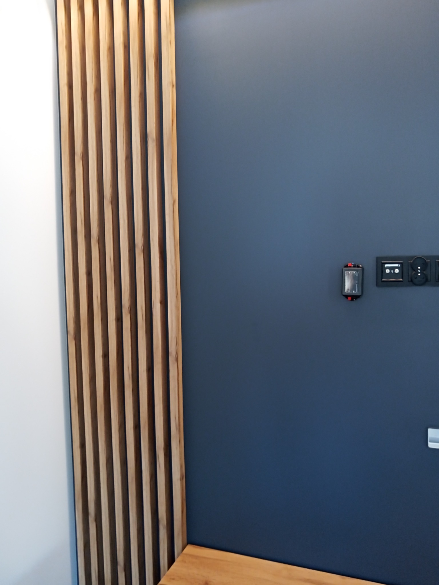 Drewniane lamele dekoracyjne na ścianie w kolorze granatowym, obok biała ściana, widoczne detale wykończenia i panel sterowania.