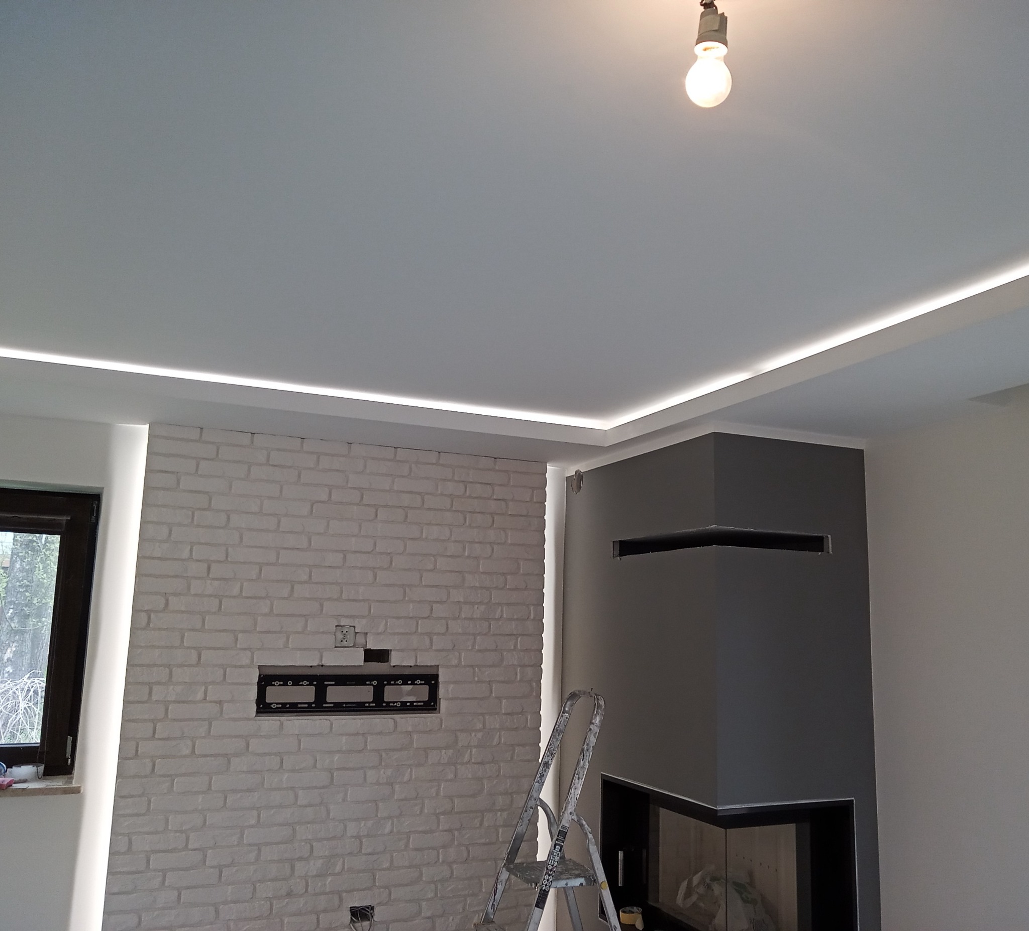 Biała ceglana ściana z zamontowanym uchwytem na telewizor, szary kominek, podwieszany sufit z oświetleniem LED, żarówka i drabina w pomieszczeniu w trakcie wykańczania.
