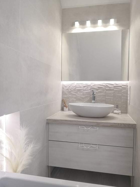 Jasna łazienka z umywalką nablatową, szafką z dwoma szufladami o nietypowych uchwytach, podświetlanym lustrem i dekoracyjną ścianą z trójwymiarowych płytek.