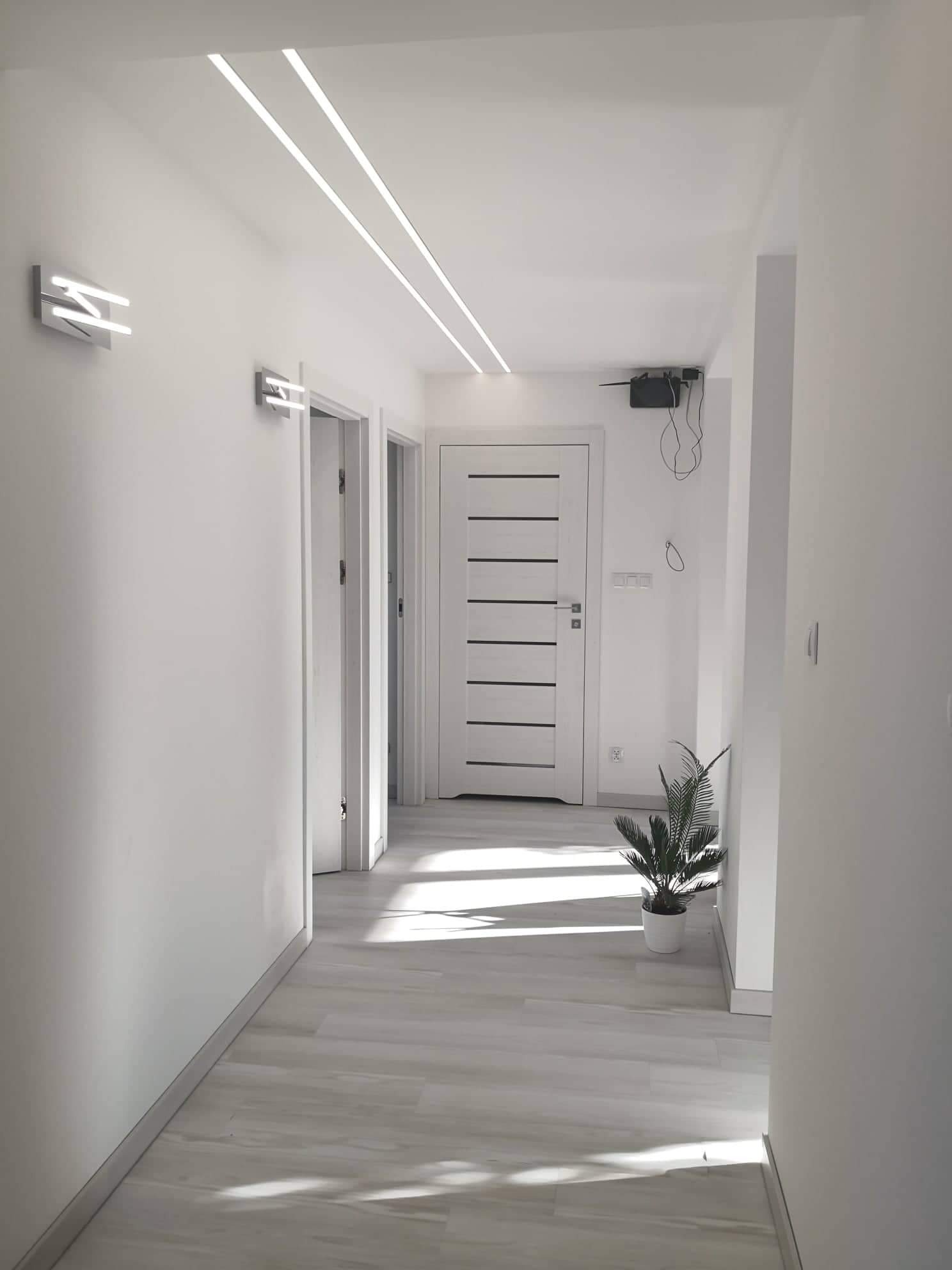 Nowoczesny korytarz w bieli z drewnopodobną podłogą, oświetleniem LED w suficie i na ścianach, oraz białą, minimalistyczną stolarką drzwiową.