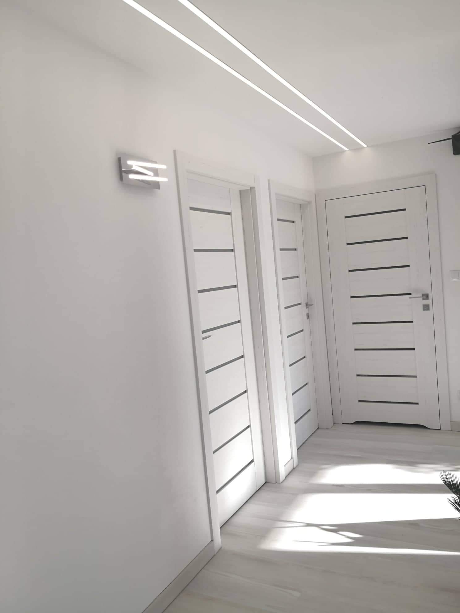 Wnętrze w bieli: trzy białe drzwi z poziomymi, ciemnymi wstawkami, oświetlenie sufitowe LED i nowoczesny kinkiet na ścianie.