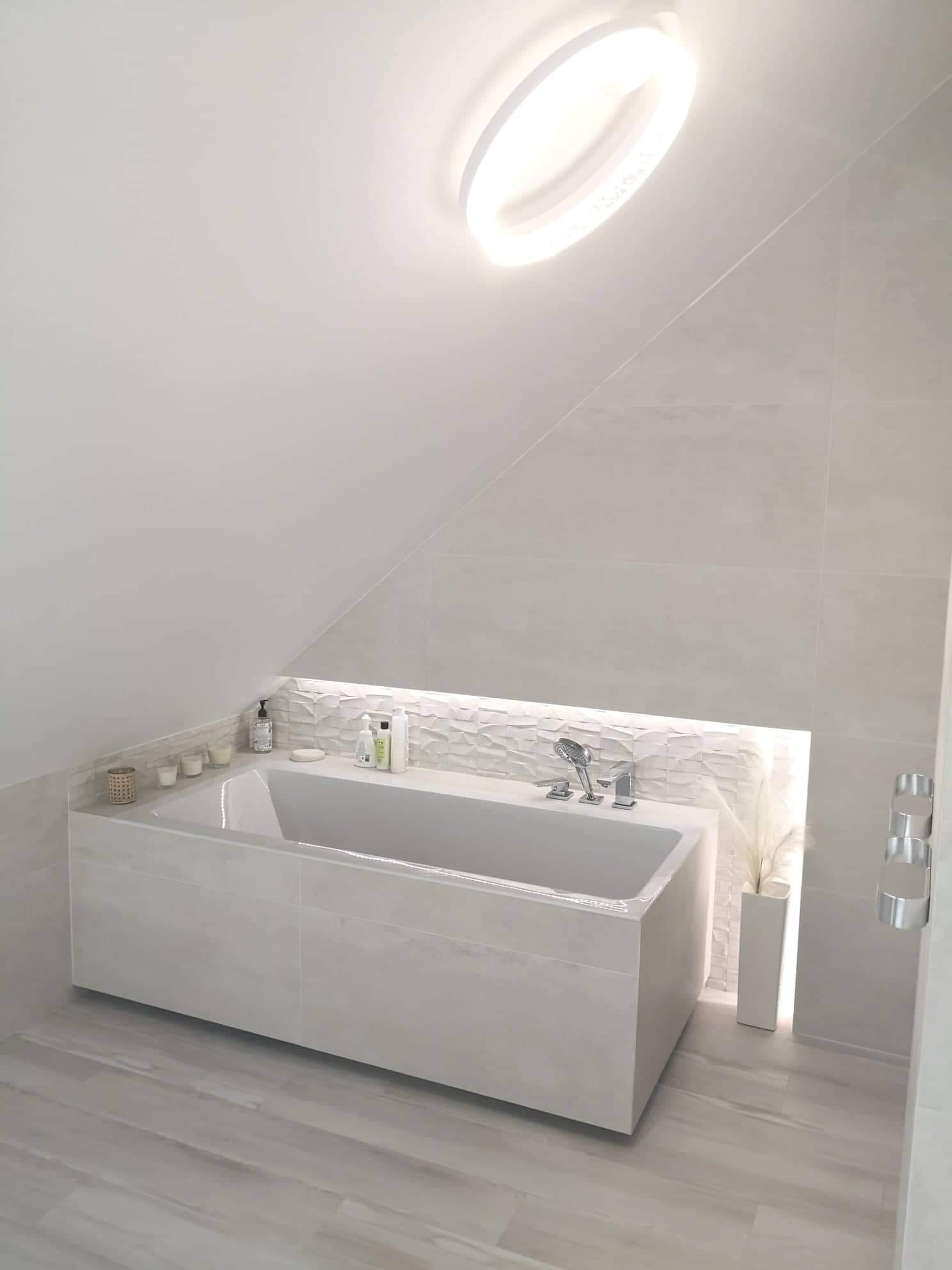 Nowoczesna, jasna łazienka na poddaszu z wolnostojącą wanną obudowaną płytkami, podświetlaną ścianą dekoracyjną i okrągłym oświetleniem sufitowym.