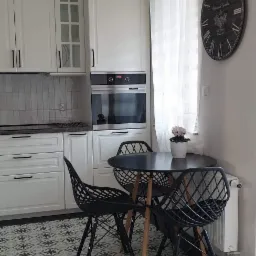 Jasna kuchnia z białymi szafkami, szarymi płytkami nad blatem, wzorzystą podłogą i czarnym okrągłym stołem z trzema krzesłami, zegar na ścianie.