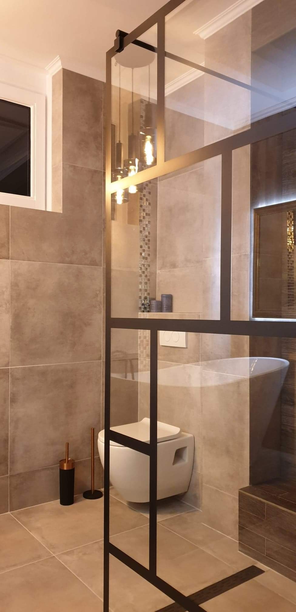 Nowoczesna łazienka z beżowymi płytkami, wiszącą toaletą i kabiną prysznicową z czarnymi profilami, oświetlona wiszącymi lampami z żarówkami Edisona.