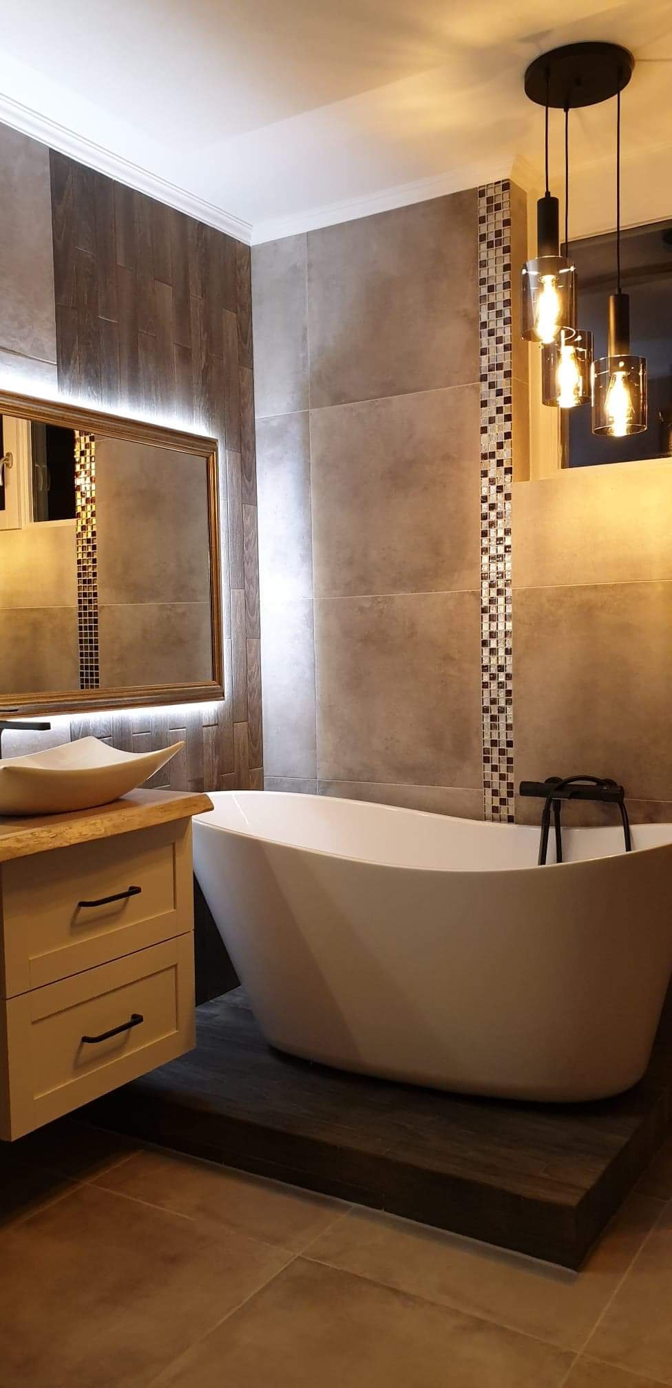 Nowoczesna łazienka z wolnostojącą wanną na podeście z ciemnego drewna, umywalką nablatową o nieregularnym kształcie, lustrem z podświetleniem LED i dekoracyjnym pasem mozaiki na ścianie.