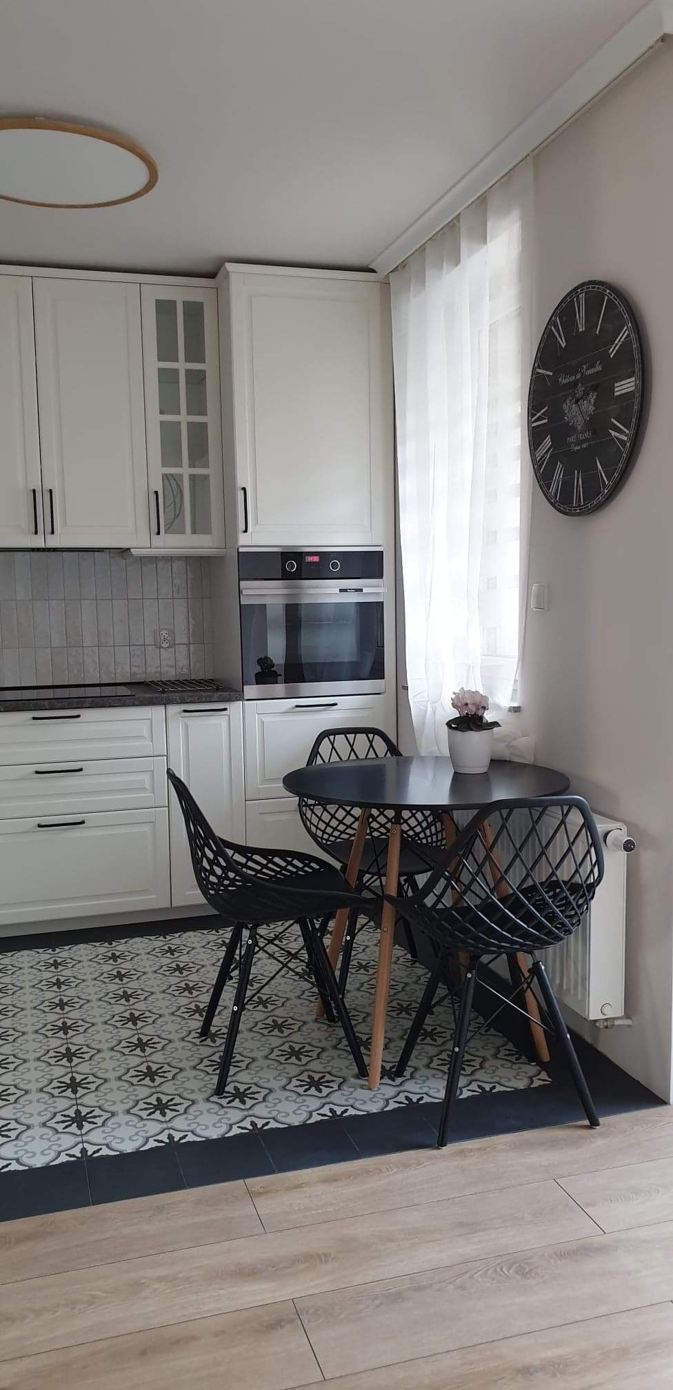 Jasna kuchnia z białymi szafkami, szarymi płytkami nad blatem, wzorzystą podłogą i czarnym okrągłym stołem z trzema krzesłami, zegar na ścianie.