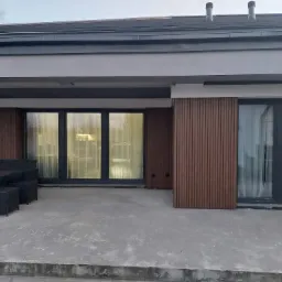 Zadaszony taras z betonową posadzką, wyposażony w czarny komplet mebli ogrodowych, z widoczną elewacją budynku wykończoną drewnopodobnymi panelami pionowymi i dużymi oknami.