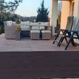 Zewnętrzny taras z desek kompozytowych w kolorze ciemnego drewna, wyposażony w meble ogrodowe z rattanu i plastikowe krzesła, oświetlony popołudniowym słońcem.