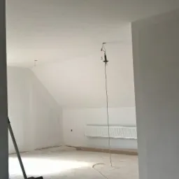 Wnętrze poddasza po malowaniu ścian na biało, widoczny grzejnik i prowizoryczne oświetlenie z żarówkami, ślady po pracach wykończeniowych na podłodze.