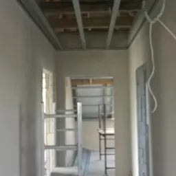 Wnętrze w trakcie montażu sufitu podwieszanego z widoczną konstrukcją metalową, drewnianym stropem powyżej, oraz rozłożonymi elementami na podłodze i rusztowaniem.