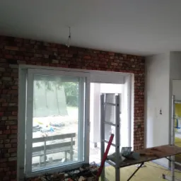 Wnętrze w trakcie remontu: ściana wyłożona cegłą klinkierową, białe okno przesuwne, drabina i st&oacute;ł roboczy z narzędziami, widoczne niedokończone prace.