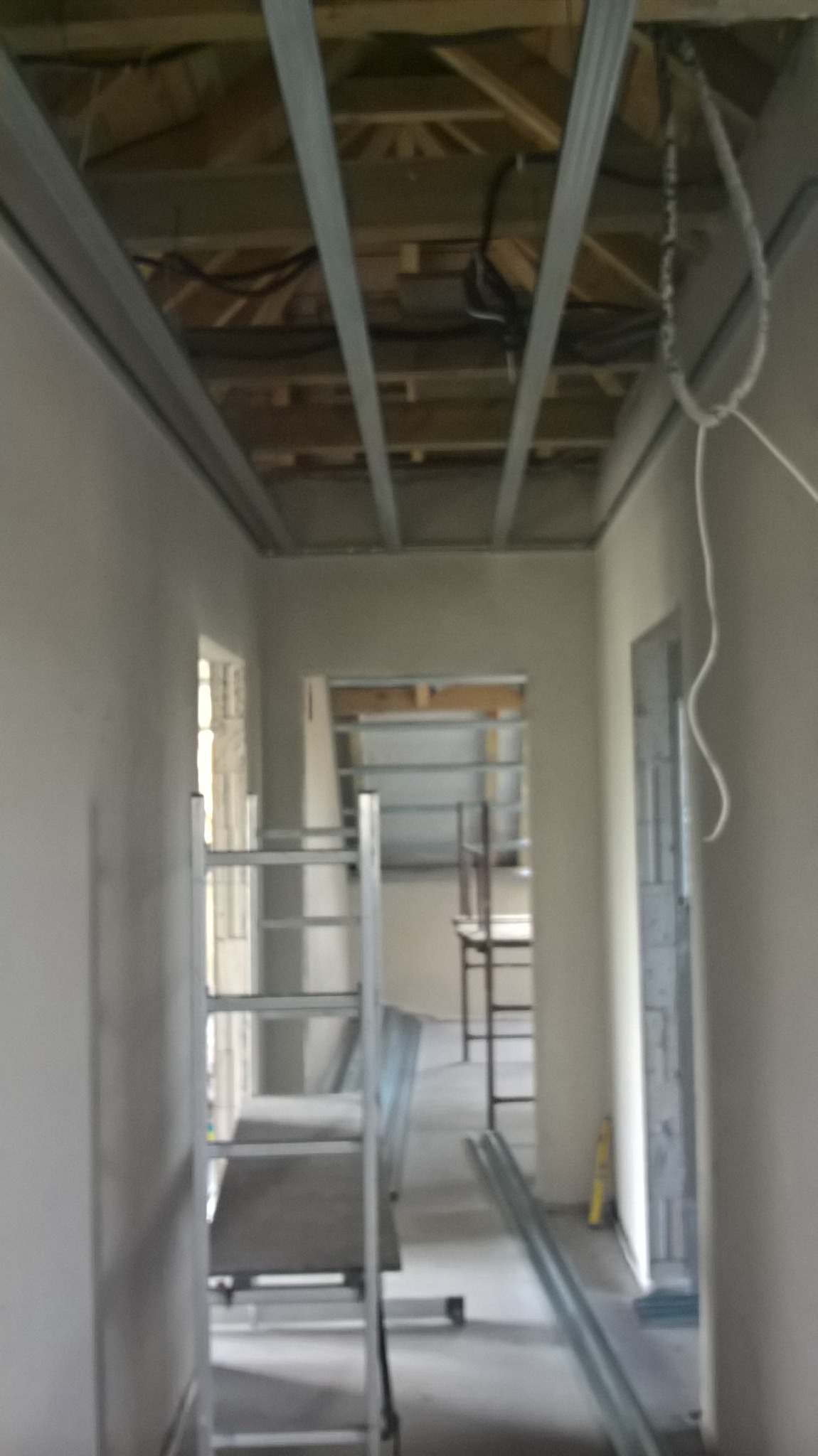 Wnętrze w trakcie montażu sufitu podwieszanego z widoczną konstrukcją metalową, drewnianym stropem powyżej, oraz rozłożonymi elementami na podłodze i rusztowaniem.