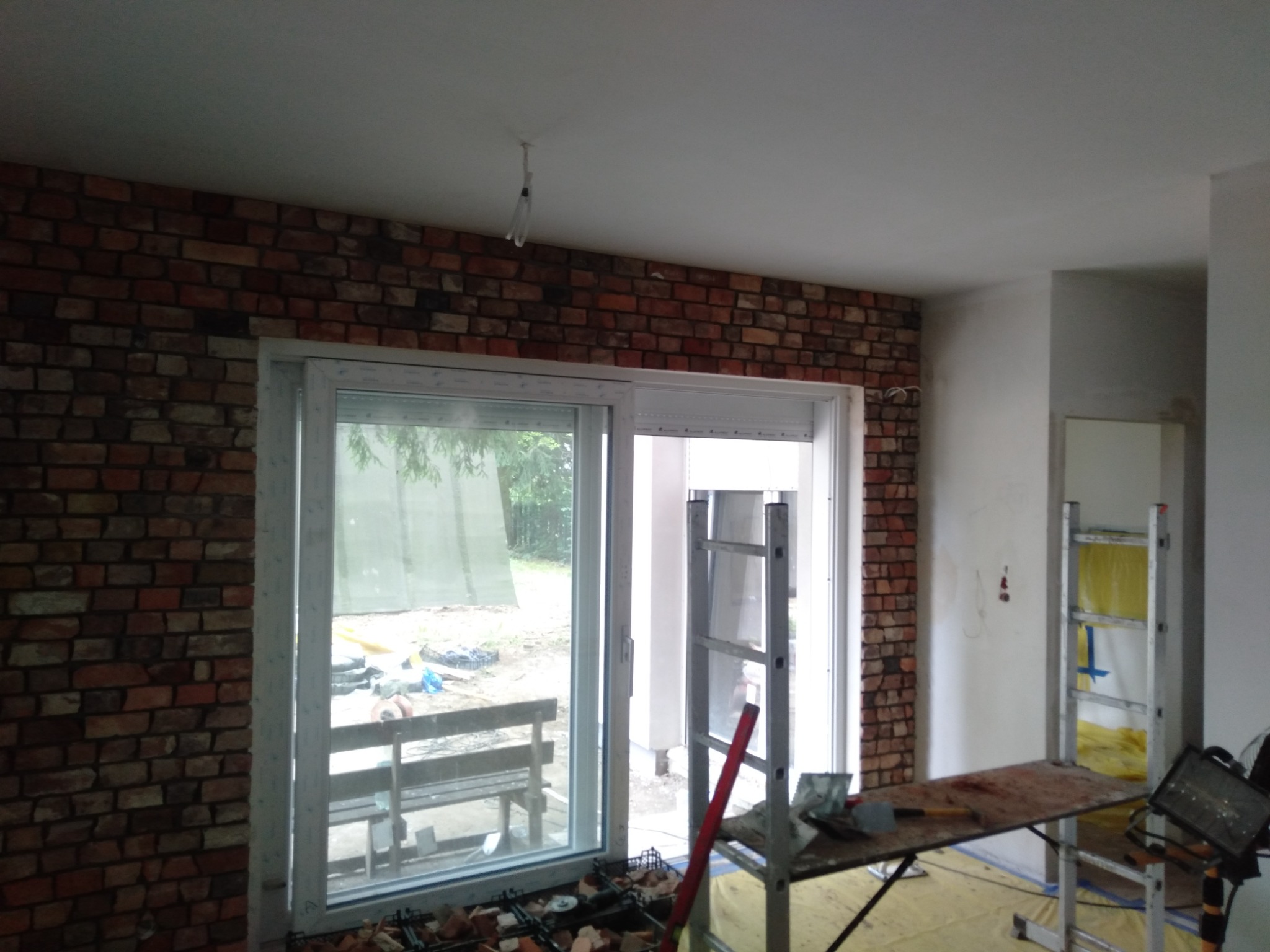 Wnętrze w trakcie remontu: ściana wyłożona cegłą klinkierową, białe okno przesuwne, drabina i stół roboczy z narzędziami, widoczne niedokończone prace.