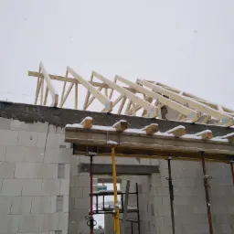 Wznoszenie ścian domu z bloczków i konstrukcji drewnianej dachu, częściowo pokrytej śniegiem, widoczne rusztowania i pochmurne niebo.