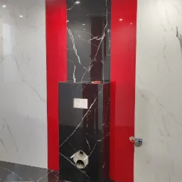 Wnętrze łazienki w trakcie wykańczania z czarno-białymi marmurkowymi płytkami na podłodze i ścianach, z widoczną instalacją podtynkową WC obudowaną czarną płytką na tle czerwonego panelu.