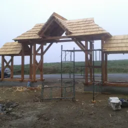 Drewniana konstrukcja z dachem pokrytym gontem, w trakcie budowy, z widocznymi rusztowaniami i materiałami budowlanymi wokół, na tle pochmurnego nieba.