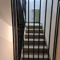 Czarne, metalowe zabezpieczenie schodów z widocznym zamkiem i uchwytem, klatka schodowa z lastryko w tle.