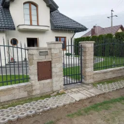 Nowoczesne ogrodzenie z metalowymi przęsłami i kamiennymi słupkami, z brązową skrzynką na listy wbudowaną w jeden ze słupków, prowadzące do domu z szarą dachówką.