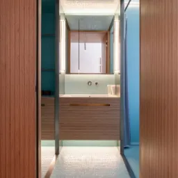 Wnętrze łazienki z mozaikową podłogą i ścianami w odcieniach turkusu, drewniane szafki pod umywalką, pionowe lampy po bokach lustra, widok z korytarza z drewnianą podłogą.