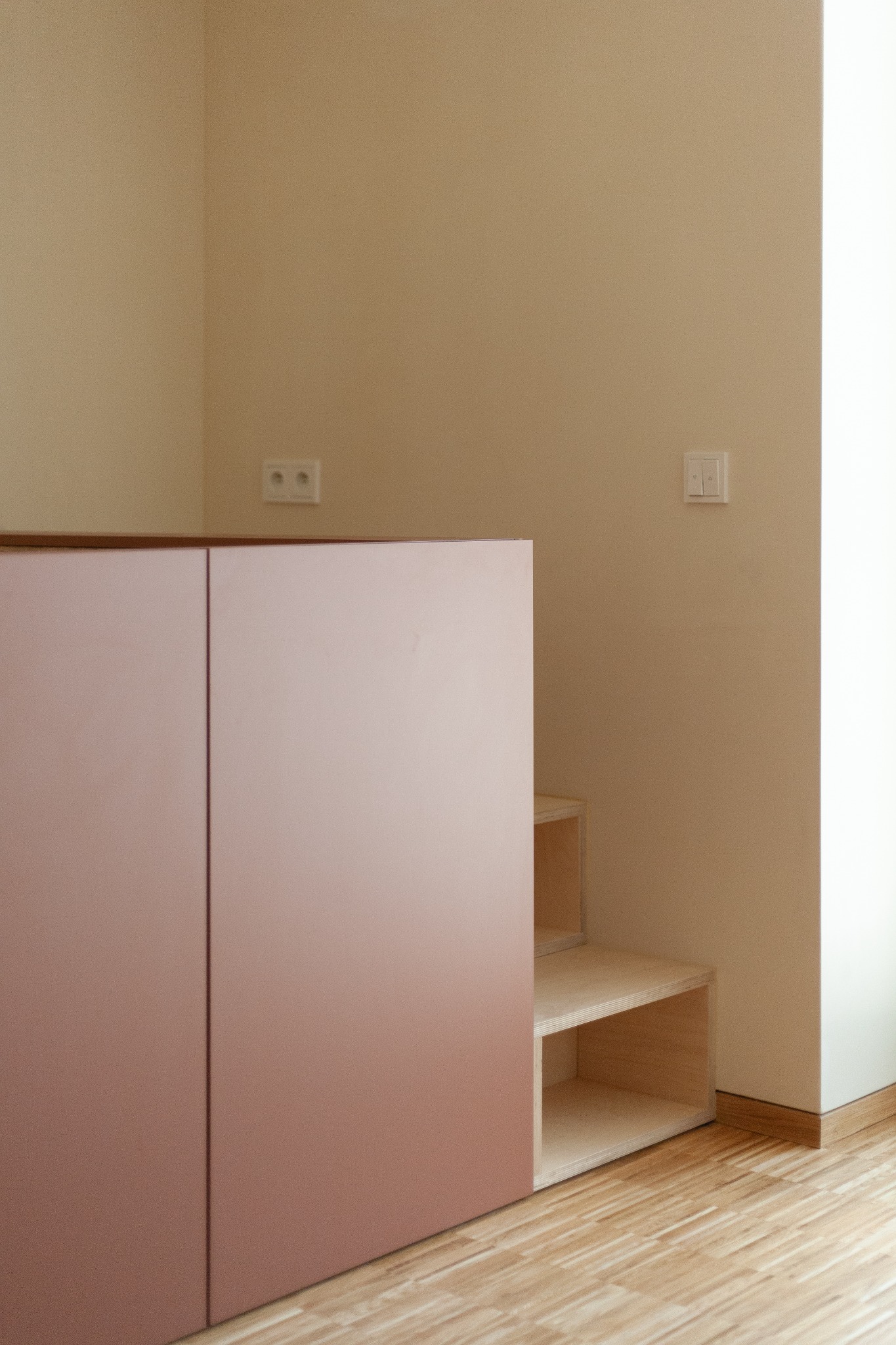 Minimalistyczne wnętrze z zabudową schodkową z jasnego drewna i różowymi frontami szafek, widoczna podłoga z paneli i gniazdko elektryczne na beżowej ścianie.