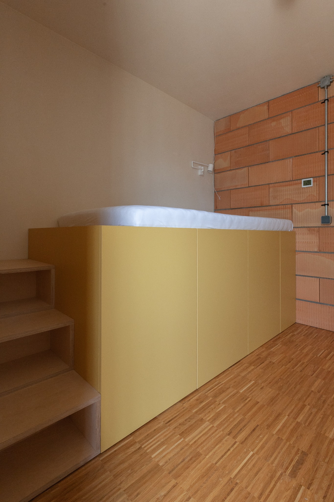 Podwyższone łóżko z żółtymi panelami i schodkami z jasnego drewna, surowa ściana z cegieł i drewniana podłoga w minimalistycznym pokoju.