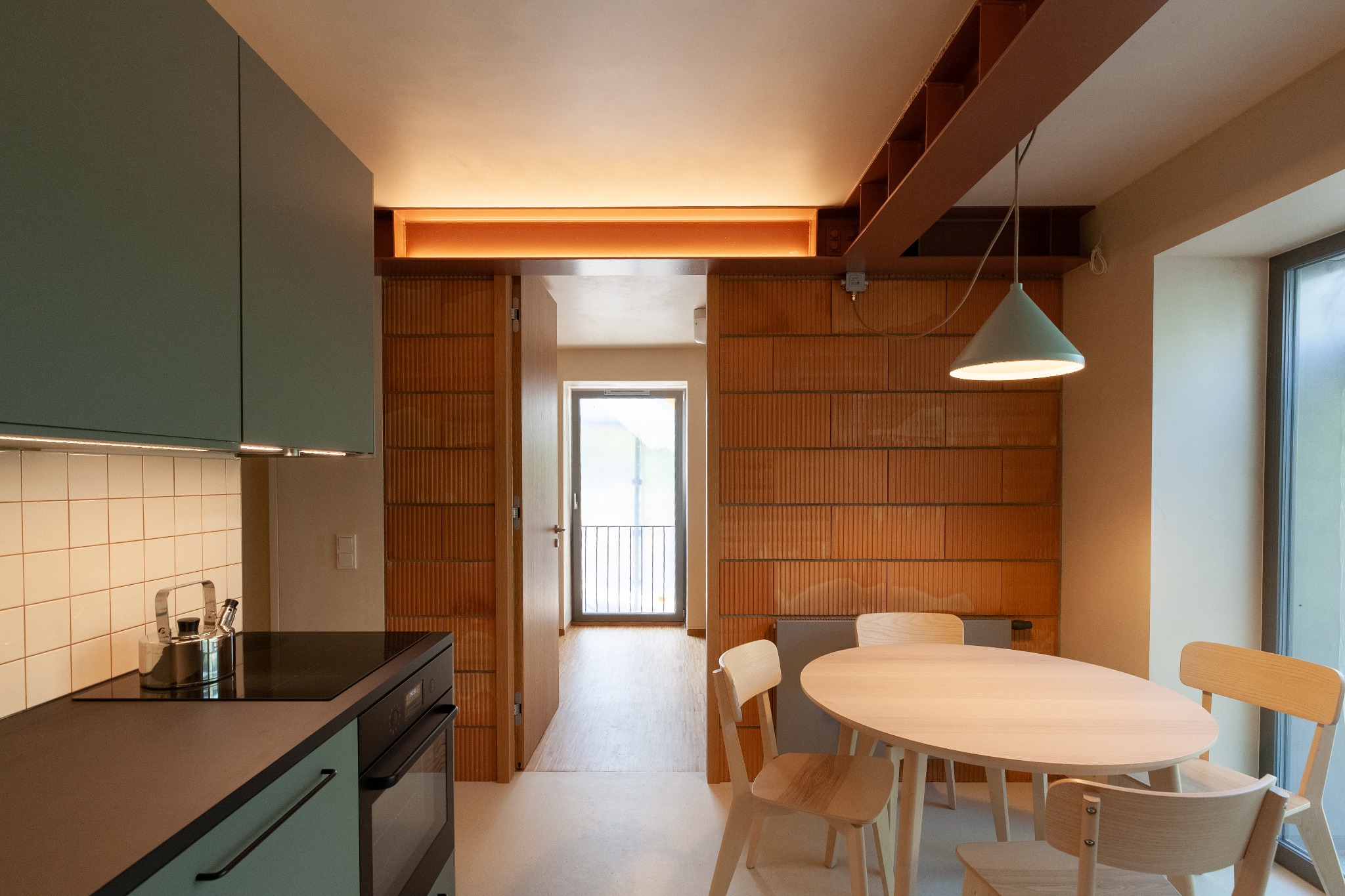 Nowoczesne wnętrze mieszkania z ceglanymi ścianami, drewnianym stołem i krzesłami, zielonymi szafkami kuchennymi oraz widokiem na balkon przez otwarte drzwi.