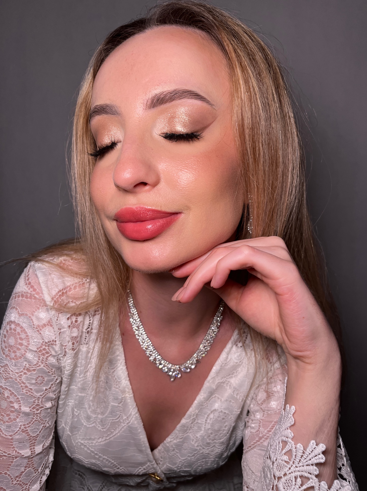 Portret kobiety z zamkniętymi oczami, prezentującej delikatny, błyszczący makijaż oczu w odcieniach złota i brzoskwini, podkreślony różową pomadką, w białej koronkowej sukience i z błyszczącym...