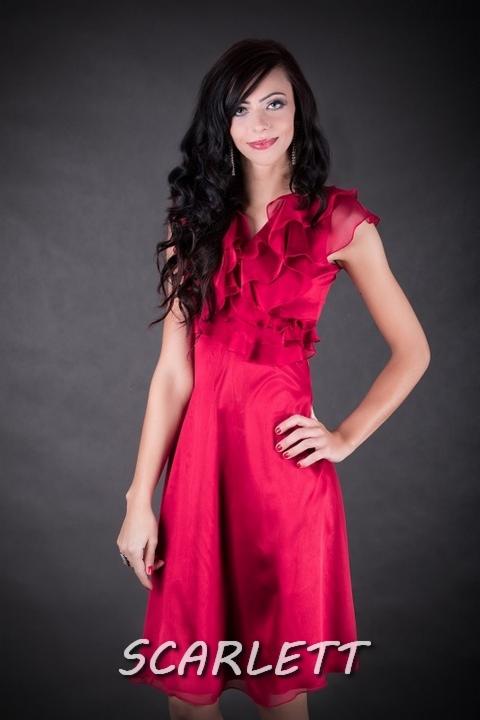 Kobieta w czerwonej sukience z falbanami pozuje na ciemnoszarym tle. Widoczny makijaż, biżuteria i delikatny uśmiech. Na dole kadru napis 'Scarlett'.