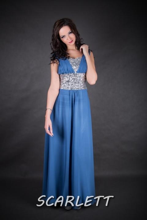 Kobieta w długiej, błękitnej sukni wieczorowej z cekinowym pasem i dekoltem, pozująca na ciemnoszarym tle. Suknia z delikatnego materiału, podkreślająca figurę.