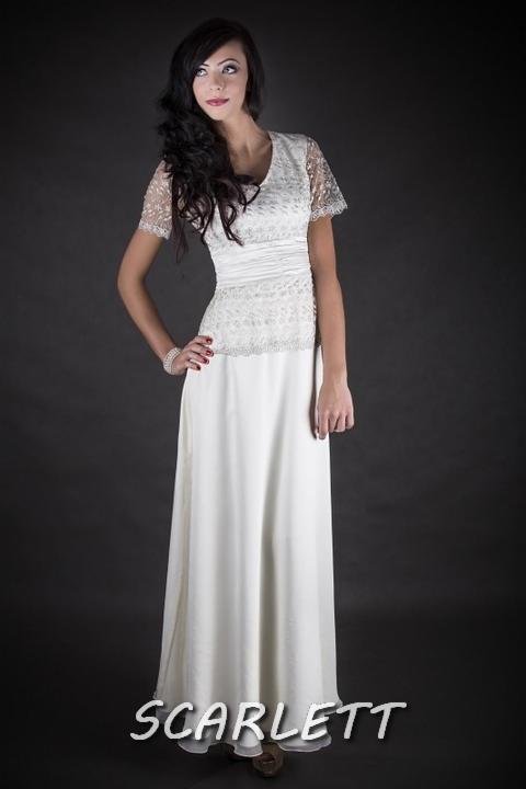 Kobieta w długiej, białej sukni z koronkową górą, pozująca na ciemnym tle. Delikatna biżuteria na nadgarstku dodaje elegancji. Subtelne oświetlenie podkreśla detale.