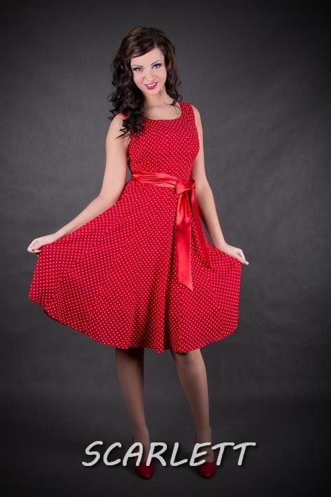 Uśmiechnięta kobieta w czerwonej sukience w białe kropki, z satynową szarfą, prezentuje kreację na ciemnoszarym tle. U dołu widoczny napis 'SCARLETT'.
